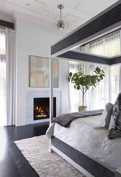 Spacious bedroom features a white and gray canopy bed on ebony floors facing a marble fireplace under art and ceiling millwork.