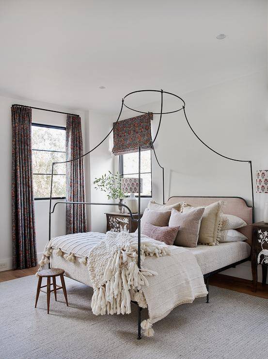 wrought iron canopy bed with a blush pink linen headboard, burlap tassel pillows, nightstands lit by white lamps.