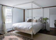 Bedroom features a white canopy bed with white bedding, a pink and beige rug layered atop a tan jute rug and a round black French bedside table.